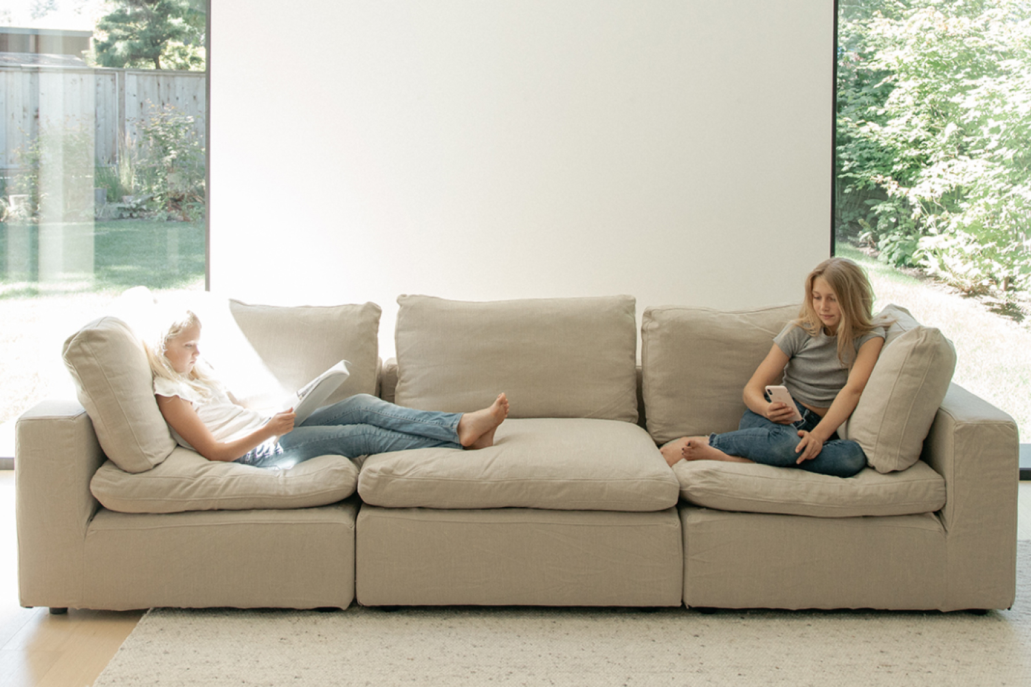 Two girls are comfortable lounging on an EM Cloud Sofa in a well-lit living room with a clear view of a green backyard through a floor-to-ceiling window.
